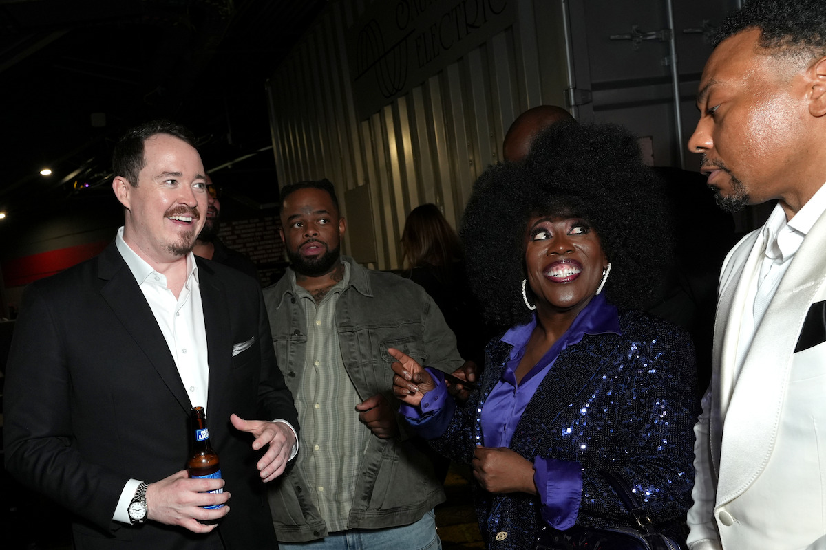 Group of people in formal and semi-casual attire socializing indoors at a nighttime event, with one woman in a sequined jacket smiling and gesturing as others listen and interact around her.