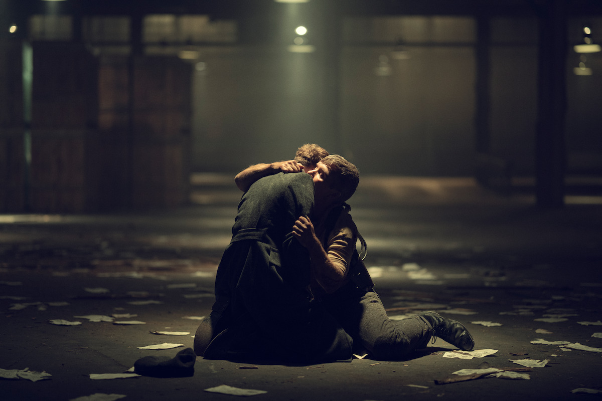 Two people kneel on the floor of a dimly lit warehouse, embracing each other, surrounded by scattered papers and crates in a dramatic, emotional setting.
