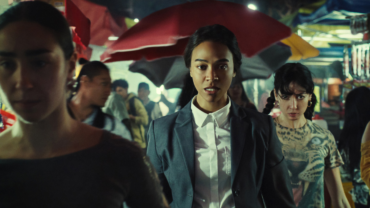 Rita (Zoe Saldaña) stands in a marketplace in a gray suit.