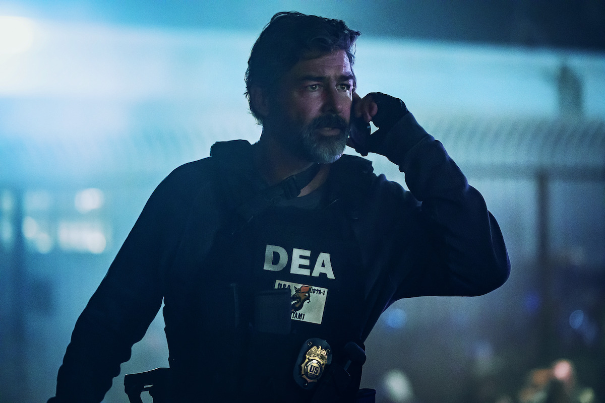 A male DEA agent with a beard and tactical vest stands outdoors at night, holding a radio to his ear. The background is dimly lit with a blue hue and blurred lights, suggesting an active scene or law enforcement operation.