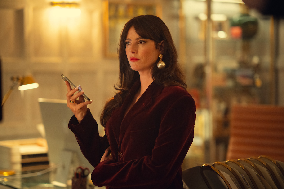 Woman in a burgundy blazer holding a smartphone stands in a warmly lit, modern office setting with bookshelves, a desk lamp, and a chair in the background.