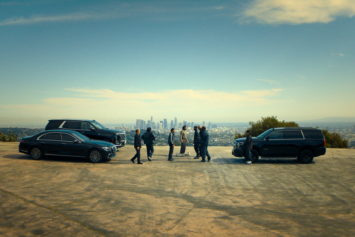 A group of people meeting between two black luxury vehicles on a hilltop overlooking a city skyline under a clear sky.