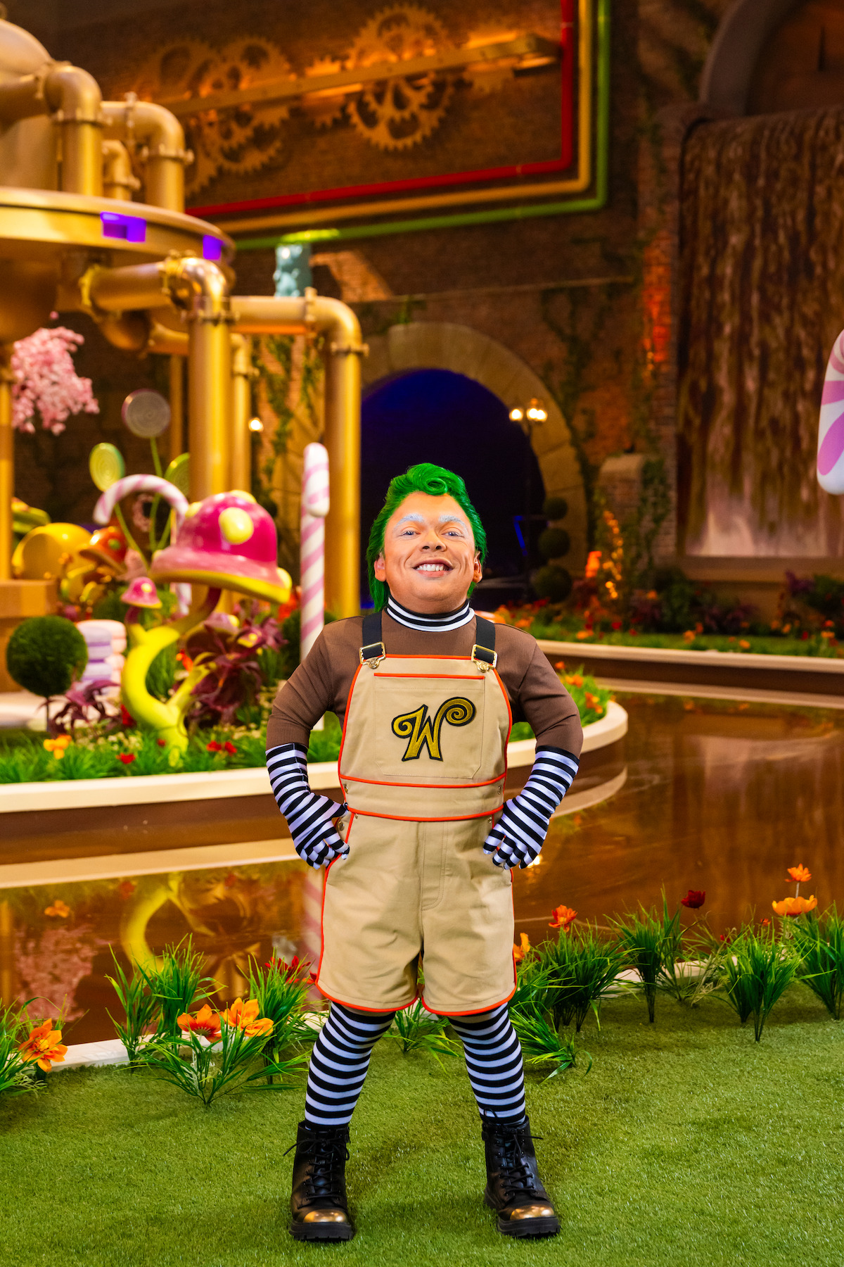 A smiling person in whimsical costume with green hair stands in a colorful, fantastical candy-themed environment featuring oversized sweets, plants, golden machinery, and a waterfall.