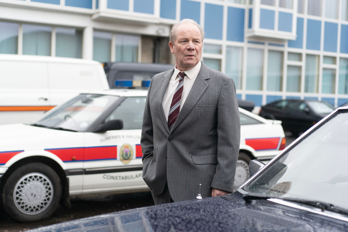 Older man in a gray suit stands in a parking lot near police cars, with a modern building in the background, appearing serious and thoughtful outdoors.