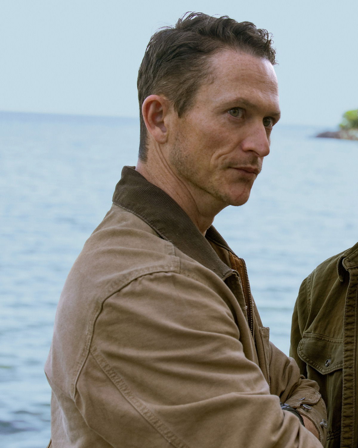Man in a tan jacket standing outdoors near a body of water, looking serious with arms crossed, partly cloudy sky and calm water in the background.