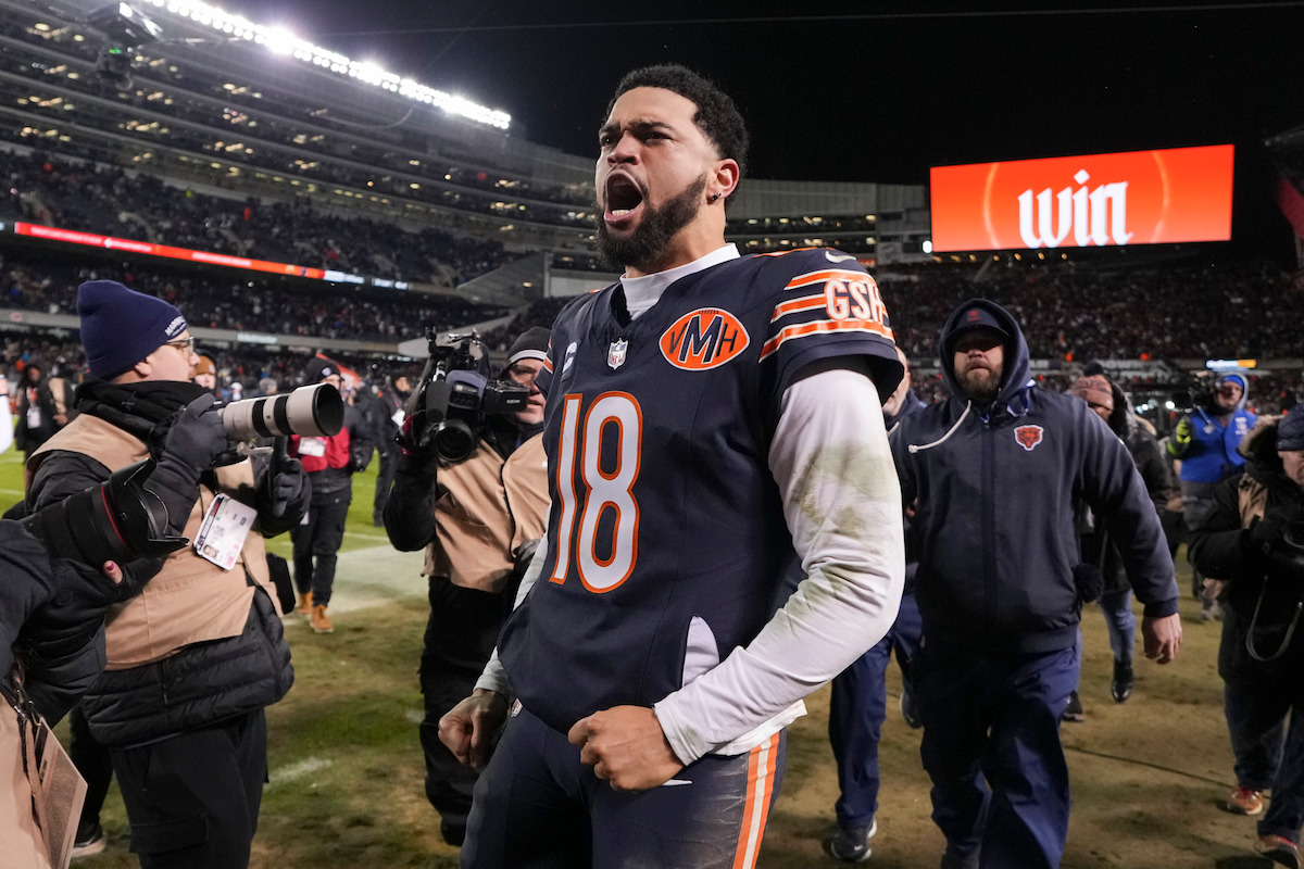 Football player celebrating on field at night, wearing Chicago Bears uniform, surrounded by photographers and team staff, stadium lights and a scoreboard reading "win" visible in the background, crowd in the stands.