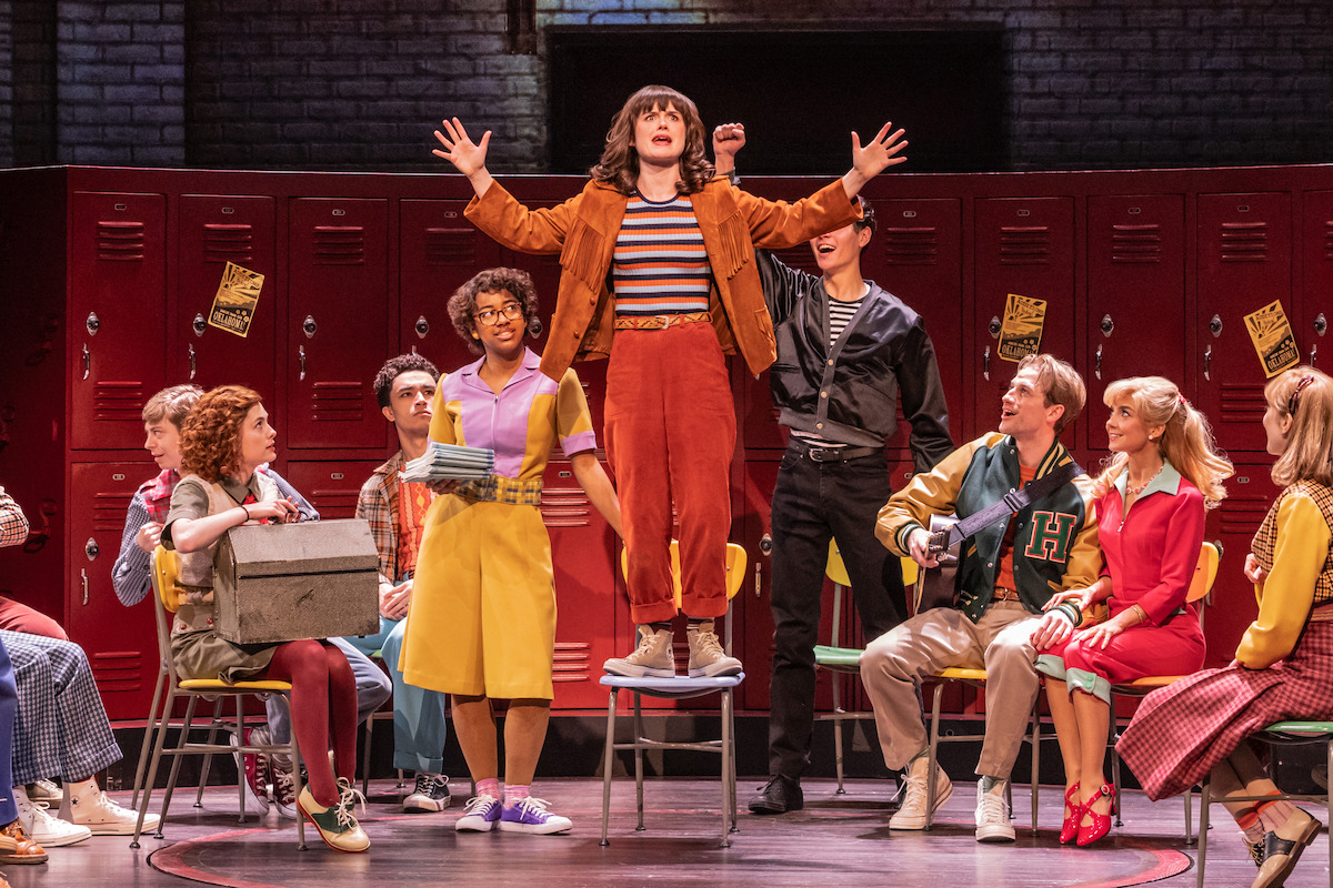Group of young actors in colorful retro costumes perform in front of red school lockers; one person stands on a chair with arms raised while others sit or interact, suggesting a lively musical or theatrical scene set in a high school.