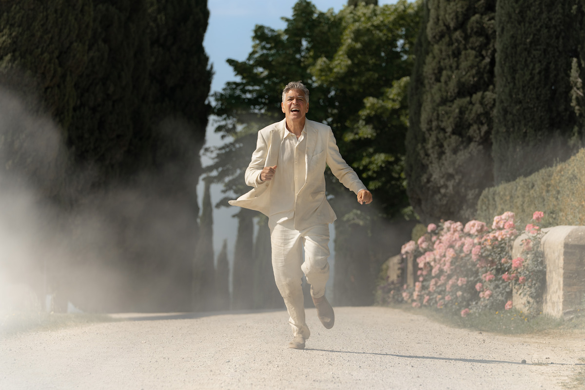 A man in a light suit runs down a tree-lined, dusty country road surrounded by flowers and greenery in bright daylight, conveying urgency and emotion in a serene, scenic environment.