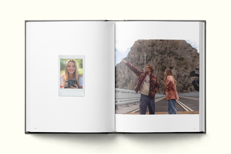 Open photo book with one page showing a woman smiling with a camera, outdoors, and the other page showing two people standing on a road pointing at a large rocky mountain in the background.