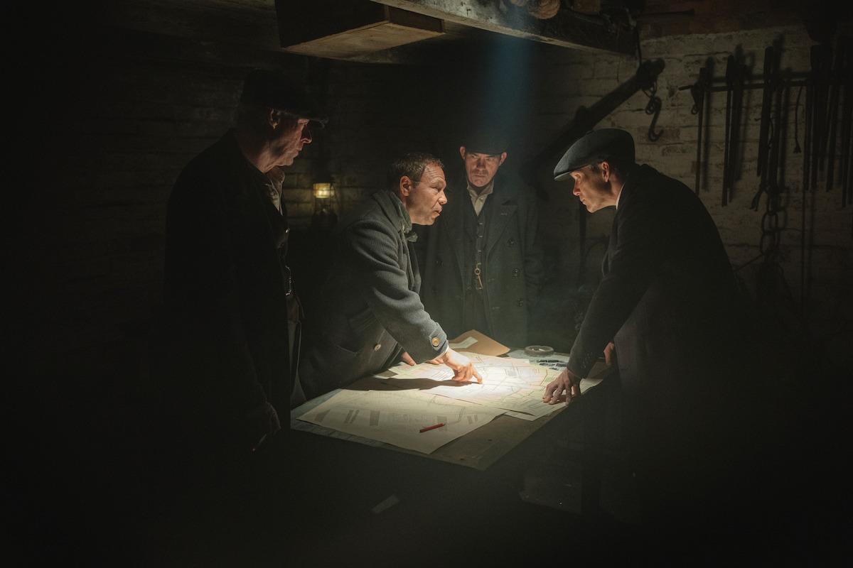 Four men in dark coats and hats gather around a large table with a map in a dimly lit, rustic room, appearing engaged in serious discussion; old tools hang on the wall, giving a vintage, secretive atmosphere.