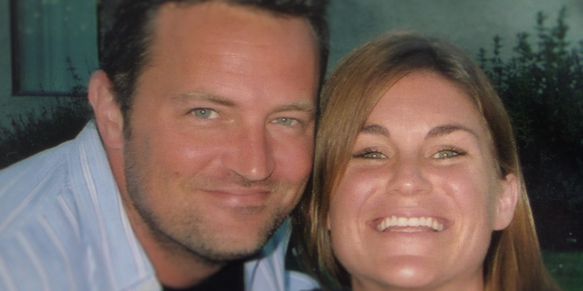 Two people smiling closely together outdoors in soft light, with greenery and a window in the background.