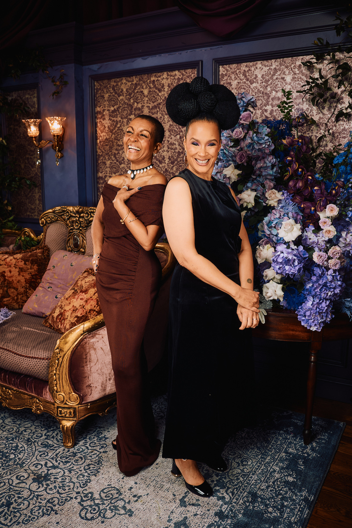 Two elegantly dressed women posing and smiling in a luxurious, vintage-style room with ornate furniture, floral arrangements, and rich blue and gold decor.
