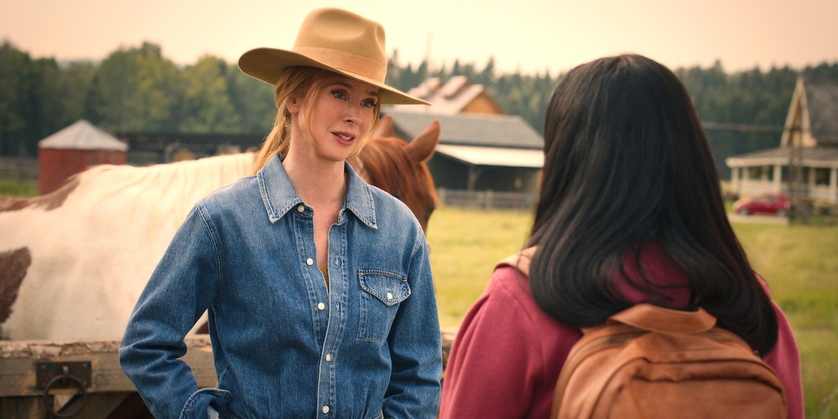 Two women talk outdoors on a ranch, one in a denim shirt and cowboy hat, the other with a backpack. A horse and farm buildings are visible in the background, with trees and a cloudy sky.