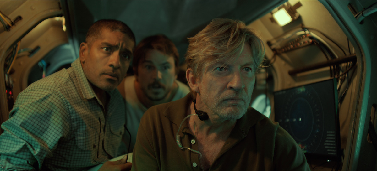 Three men in a dimly lit submarine interior look intently at a screen, surrounded by equipment and controls, suggesting a tense or serious moment in an underwater or scientific environment.