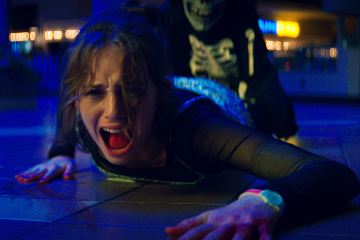 A distressed woman screaming on a tiled floor in a brightly lit indoor setting, with a person in a skeleton costume looming behind her, creating a tense and dramatic atmosphere.