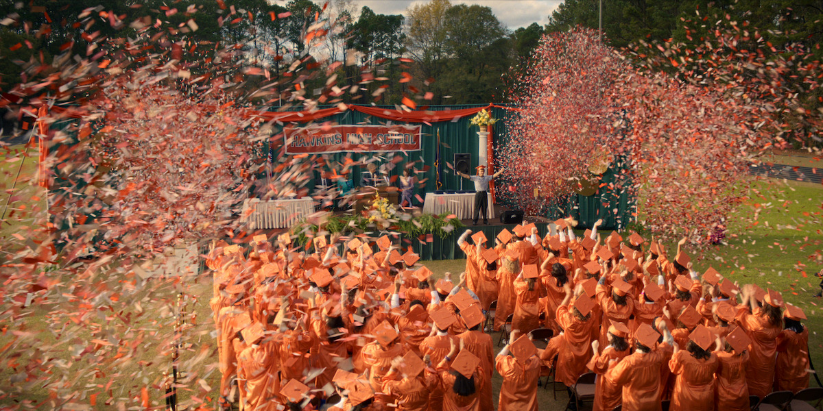 Graduates in orange caps and gowns celebrate at an outdoor graduation ceremony with confetti flying and a decorated stage in the background.