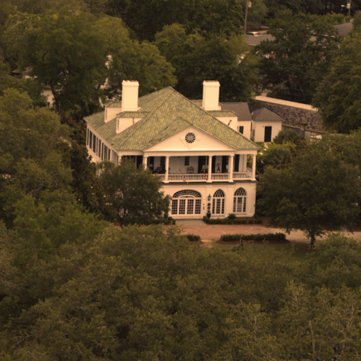 Casa branca e histórica com teto verde, duas chaminés e uma varanda cercada de árvores densas e muitas plantas em um ambiente suburbano dos EUA, visto de cima em um dia nublado.