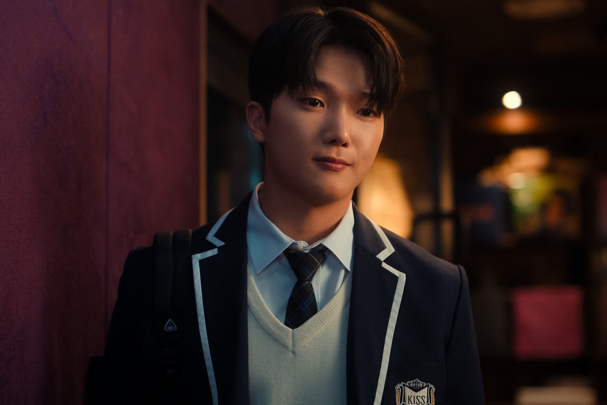 Young man in a navy school uniform with a backpack stands indoors against a purple wall, warmly lit background with blurred objects, creating a calm and studious atmosphere.