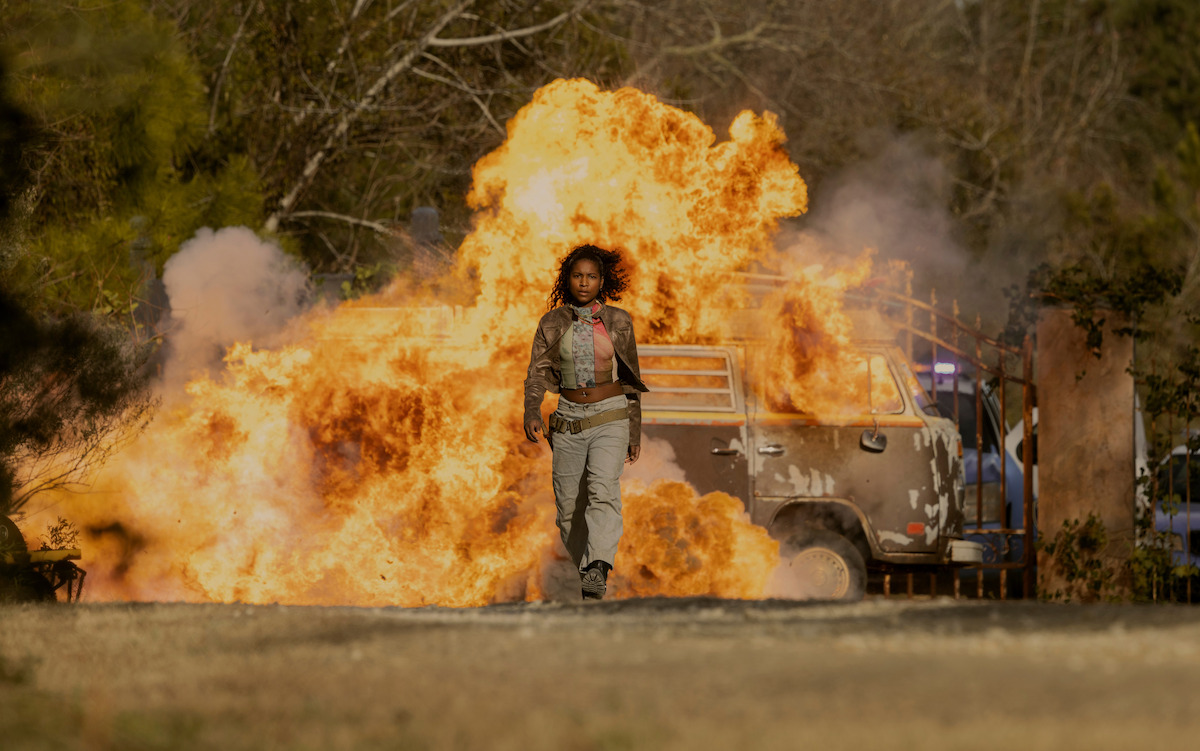 A woman walks away from a van exploding in flames outdoors, surrounded by trees and a dirt road, looking calm and determined amid a dramatic action scene.