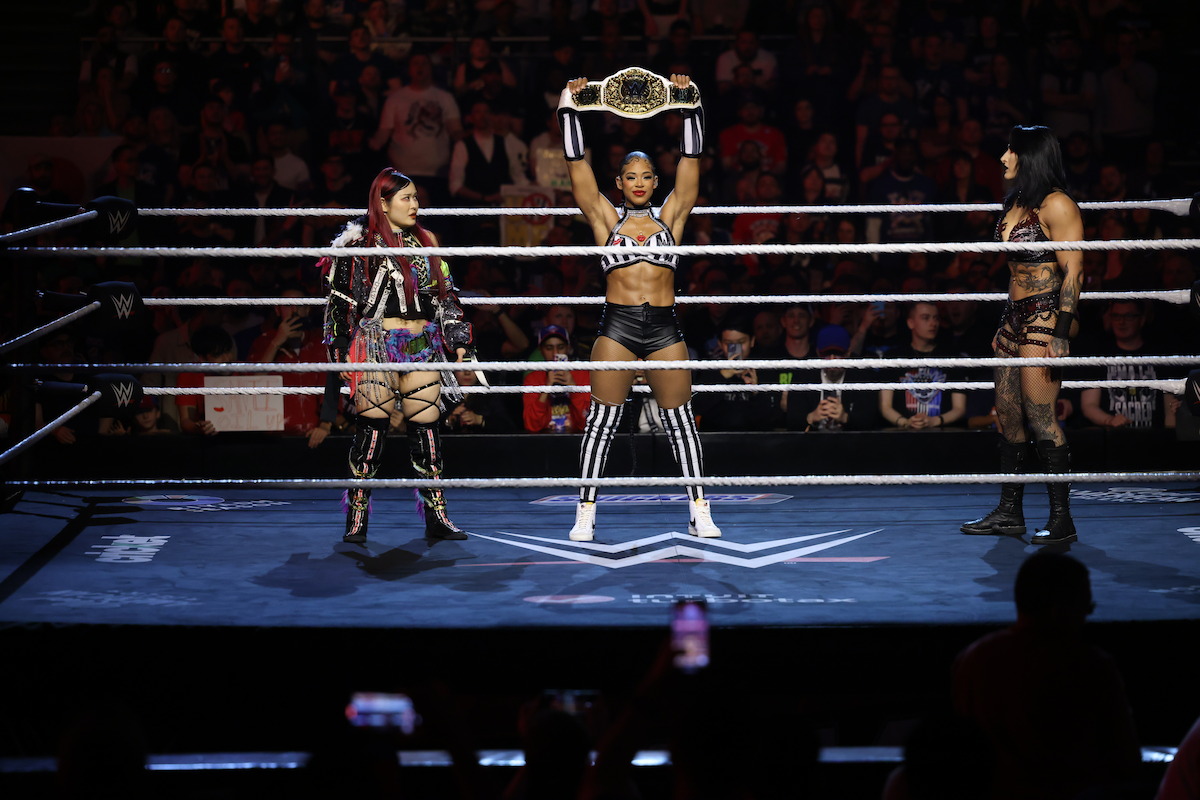 Bianca Belair holding up a belt flanked by IYO SKY and Rhea Ripley.