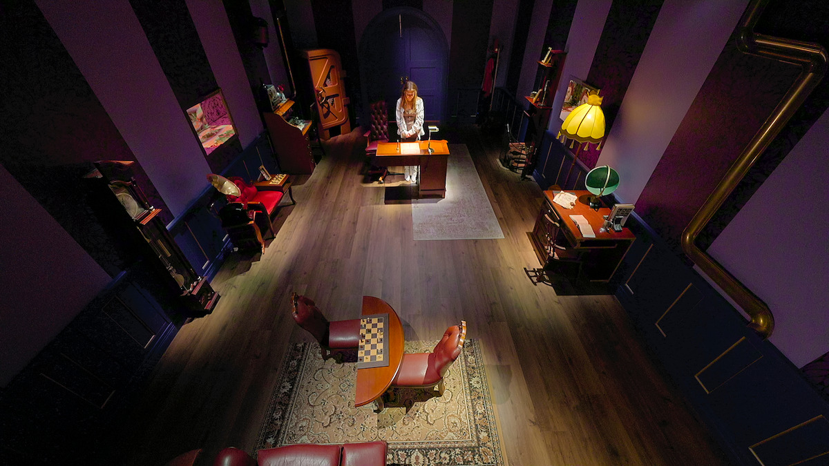 Woman standing at a desk in a stylish, vintage room with purple walls, wooden floor, chessboard table, leather chairs, bookshelves, lamps, and cozy decor, viewed from above.