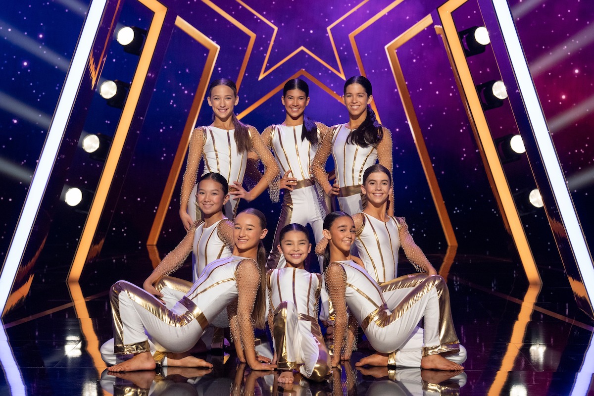 The Force in matching white and gold costumes pose together on a brightly lit stage with a star-studded background and geometric light designs.