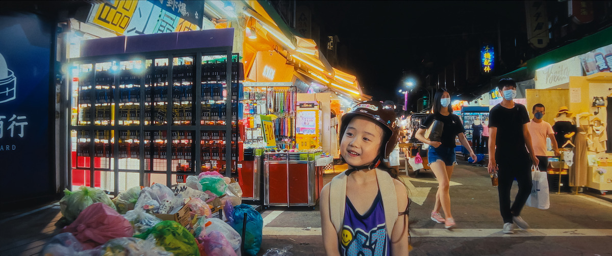Smiling child in helmet at a lively night market, surrounded by colorful lights, stalls, garbage bags, and people walking, creating a vibrant, busy urban atmosphere.