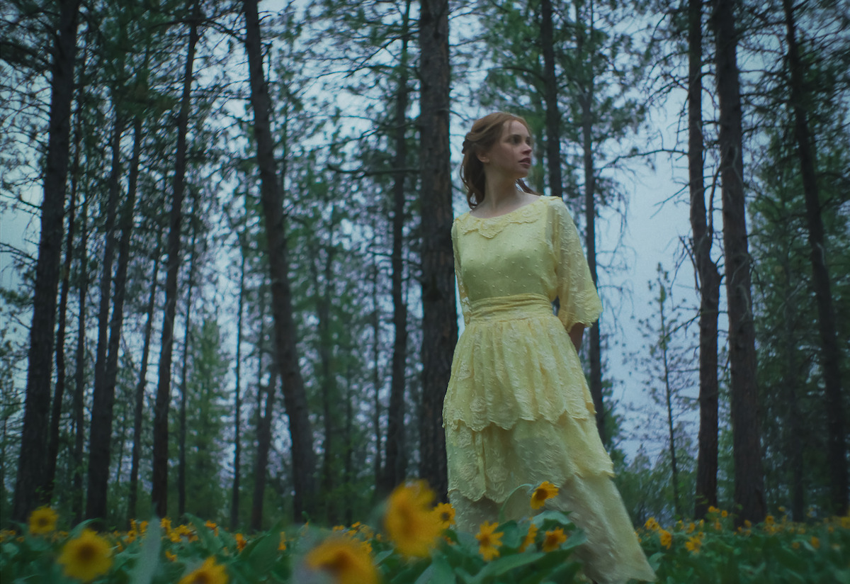 Mujer con un vestido amarillo claro parada en un bosque iluminado por el sol rodeada de altos árboles y flores silvestres, mirando pensativamente hacia un lado, evocando un estado de ánimo sereno y soñador.