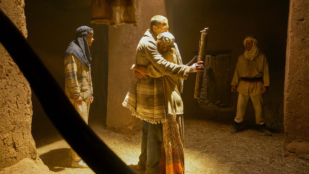 Four people in traditional clothing stand in an earthen, dimly lit room; two embrace warmly in the center while the other two look on, evoking a sense of reunion or forgiveness in a historical or biblical setting.