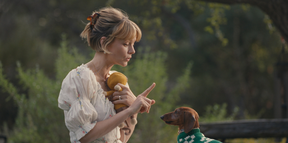 Woman in a light dress holding a toy, raising her finger while looking at a small dog wearing a green sweater outdoors in a natural, leafy setting.