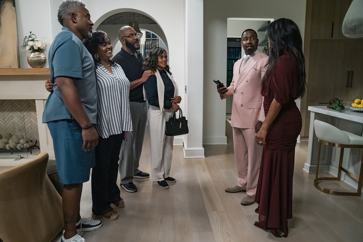 Six adults stand in a modern, well-lit living room with hardwood floors and kitchen in the background, four facing a couple dressed formally, engaged in conversation, creating a warm, social atmosphere.
