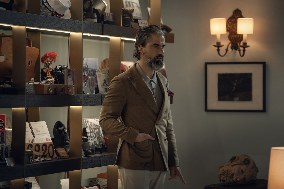 Man with gray hair and beard in a brown blazer stands in a stylish room with shelves of eclectic objects, warm lighting, wall art, and a table with a skull.