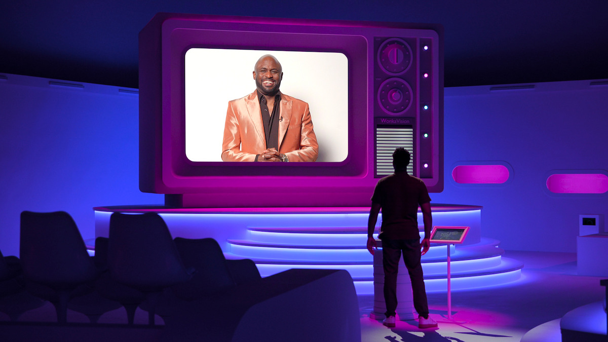 A person stands in a futuristic purple-lit room, facing a large retro-styled TV screen displaying a man in a shiny orange suit smiling against a white background. Modern seating and glowing accents fill the setting.