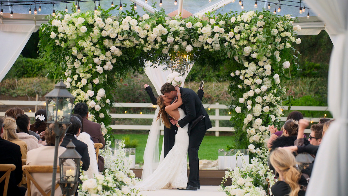 Bride and groom share a kiss under a large floral arch at an outdoor wedding ceremony, surrounded by guests seated on both sides, lush greenery, and elegant white flower arrangements.