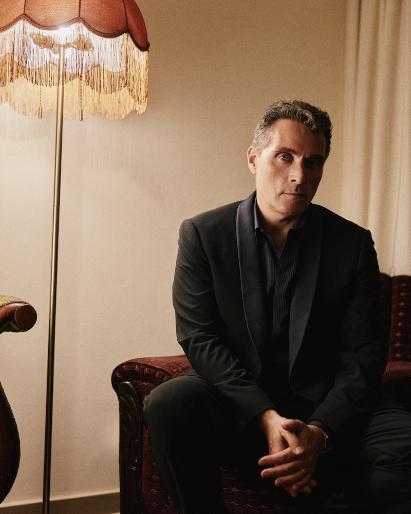 Man in a dark suit sits on a vintage sofa in a dimly lit room with beige walls, next to a retro fringed floor lamp, creating a moody, contemplative atmosphere.
