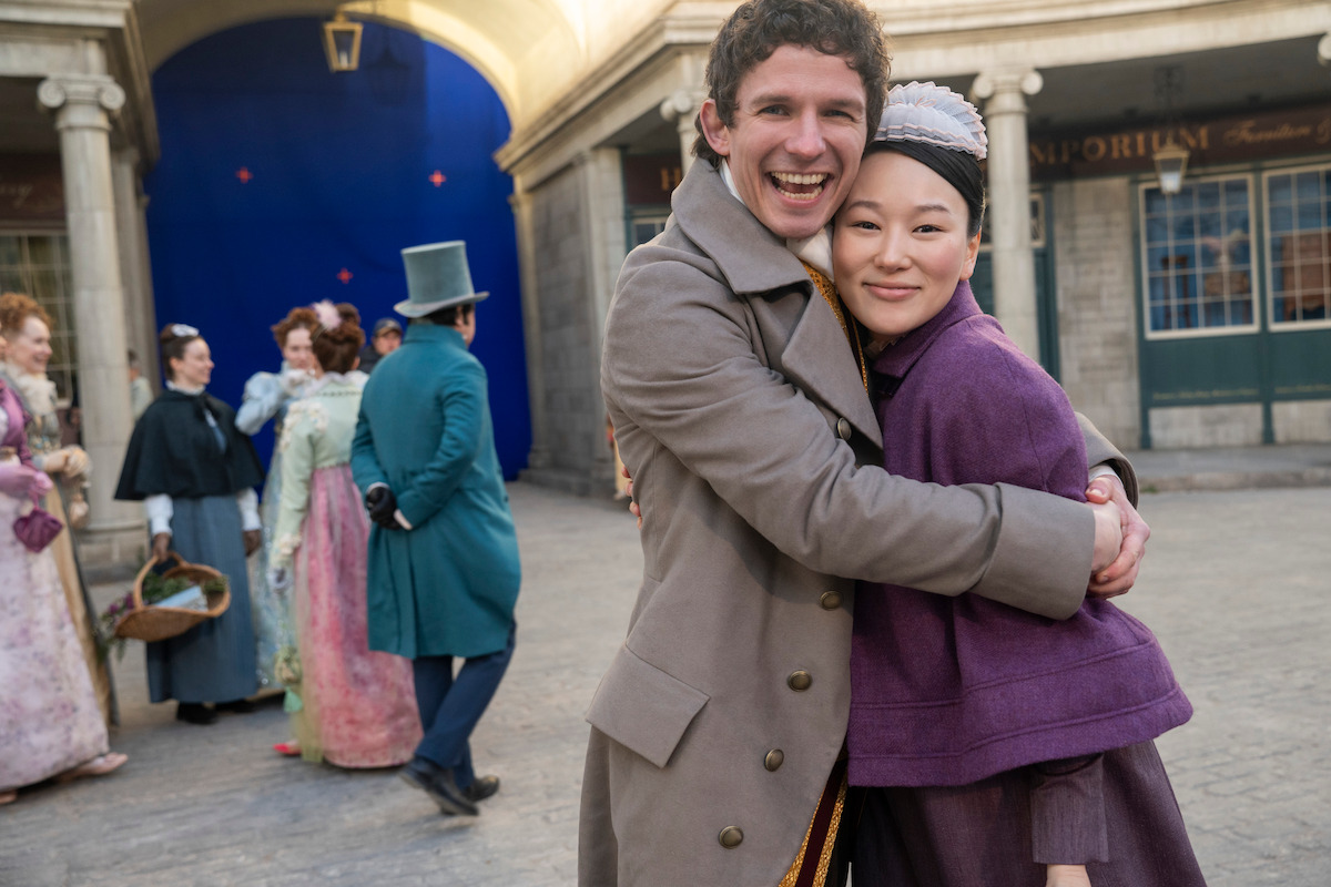 Two people in period costumes embrace and smile in a historical street setting, with other people in similar attire talking and walking in the background.