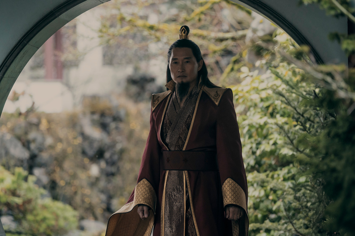 Daniel Dae Kim as Fire Lord Lord Ozai in ornate traditional robes stands in a circular garden doorway, surrounded by lush greenery and blurred natural outdoor scenery, suggesting an ancient or historical setting.