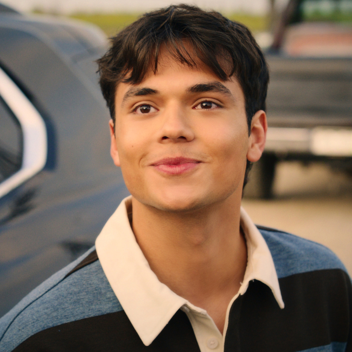 Young man smiling outdoors near a parked vehicle, wearing a blue and black striped collared shirt, with a blurred background suggesting a casual, possibly suburban or parking lot setting during daytime.
