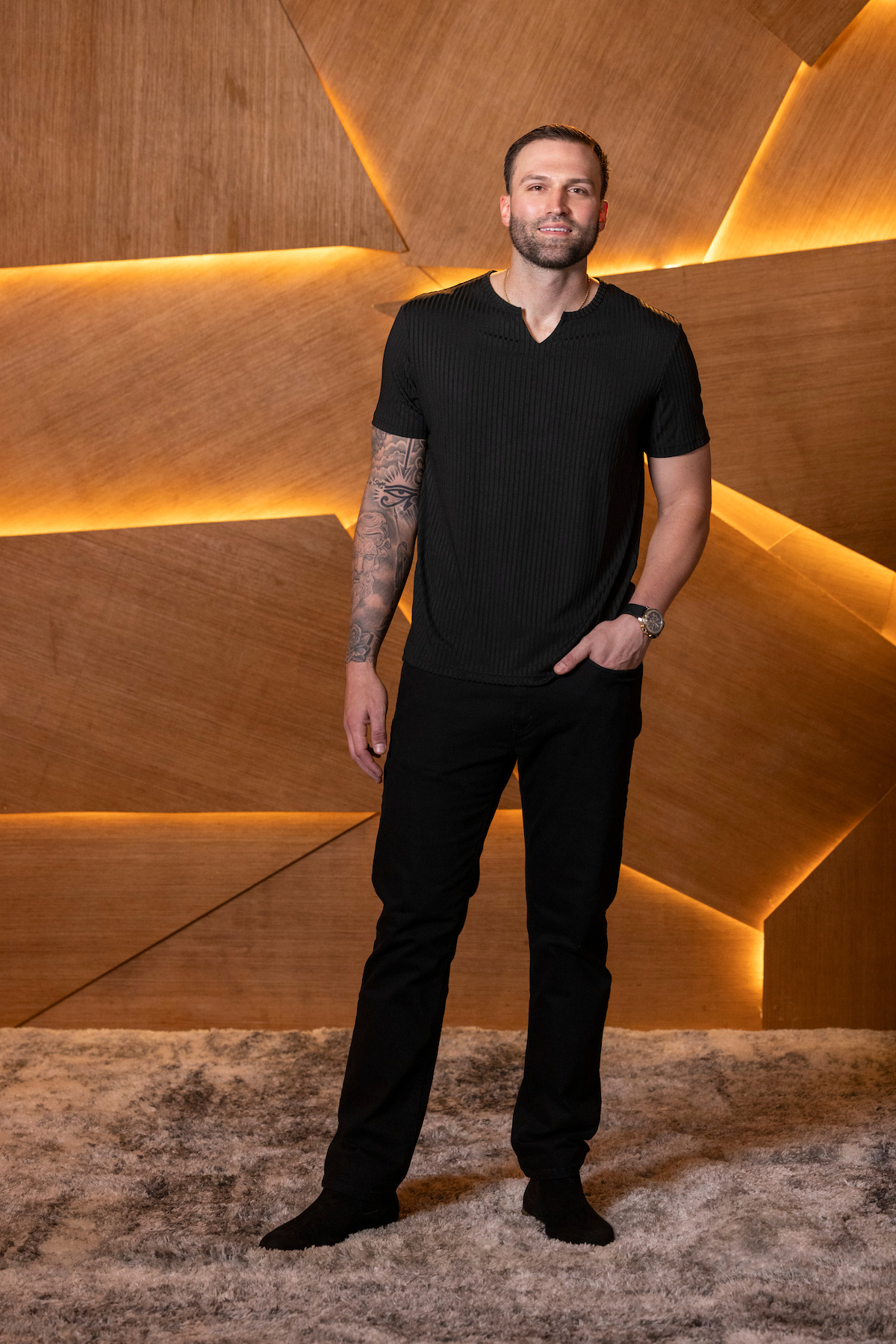 A man stands on a plush rug in front of a wooden geometric wall with glowing backlights. He wears a black shirt and pants, showing a tattooed arm and watch, in a modern, stylish indoor setting.