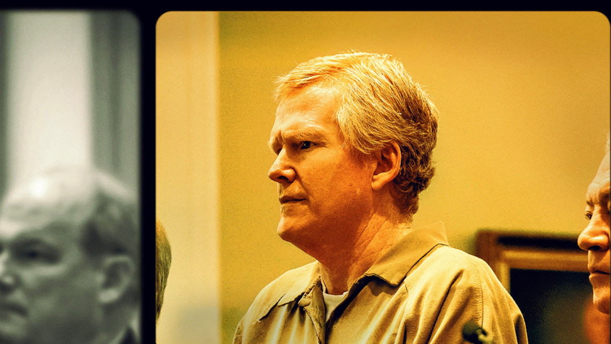 Middle-aged man with light hair in a tan prison uniform stands in a courtroom setting, with another man beside him and blurred figures in the background, all under warm indoor lighting.