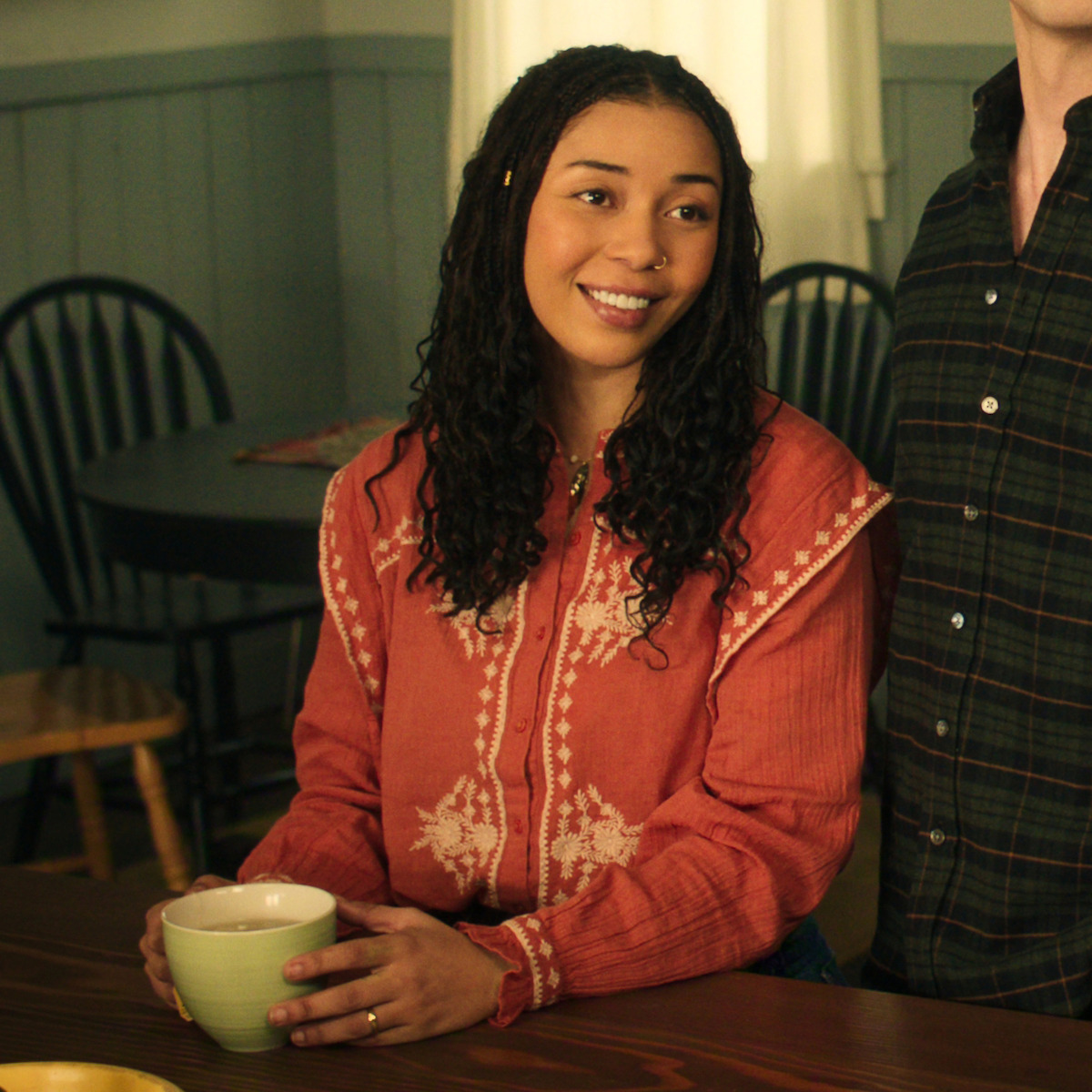 Woman with curly hair in an orange patterned shirt sits at a wooden table holding a mug, smiling, in a cozy kitchen with wooden chairs and a round table in the background. Warm, inviting atmosphere.