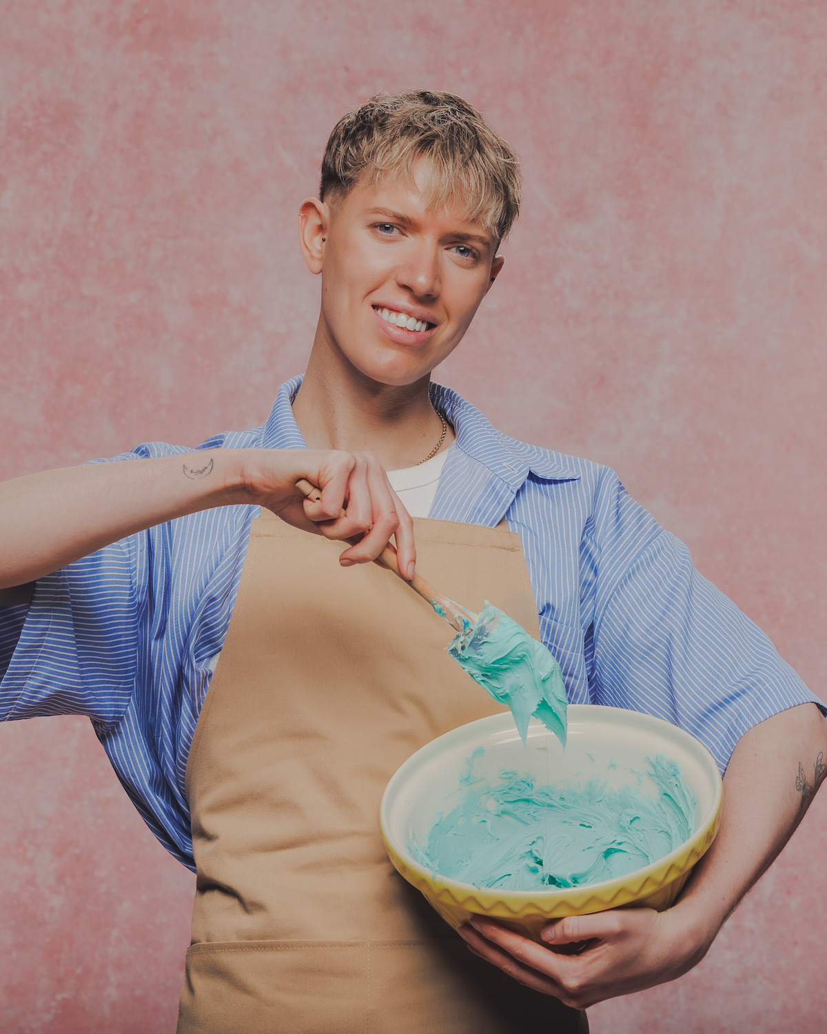 Person in tan apron and blue shirt smiling, holding a yellow mixing bowl with blue frosting against a textured pink background. Indoor studio setting with soft, diffused lighting.