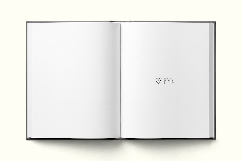 An open blank book on a light surface, with the right page showing handwritten text that says "♥ P4L" in black ink.