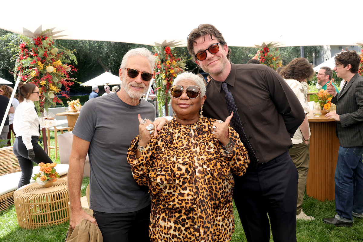 Three people posing and smiling at an outdoor event with floral decorations, tables, and other guests in the background on a sunny day.