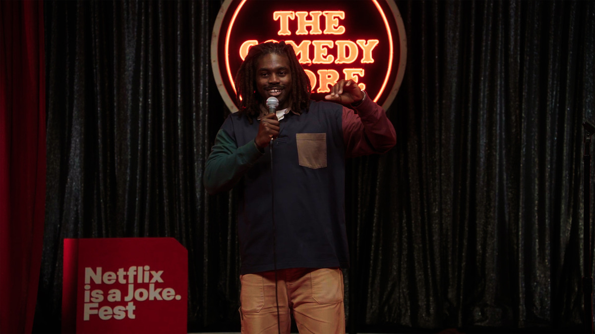 A comedian performs on stage at The Comedy Store, holding a microphone, with a neon venue sign and black curtains in the background, and a red sign reading "Netflix is a Joke Fest" to the side.