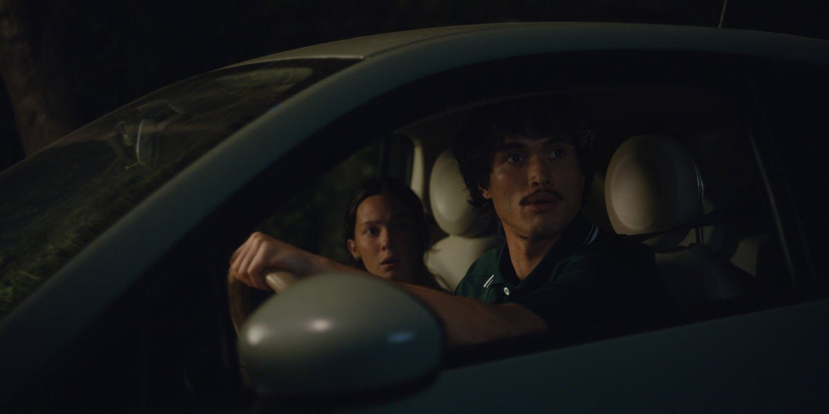 Two people sit in the front seats of a car at night, looking out the window with concerned expressions, illuminated by dim light and surrounded by darkness outside.