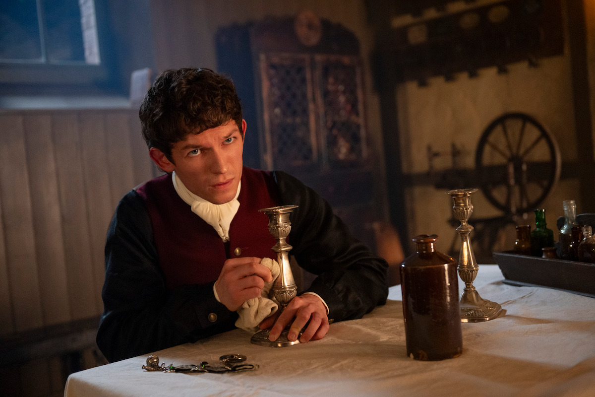 Young man in historical clothing sits at a table polishing a candlestick in a dimly lit, old-fashioned room with bottles, keys, and wooden furnishings, evoking a vintage or period atmosphere.