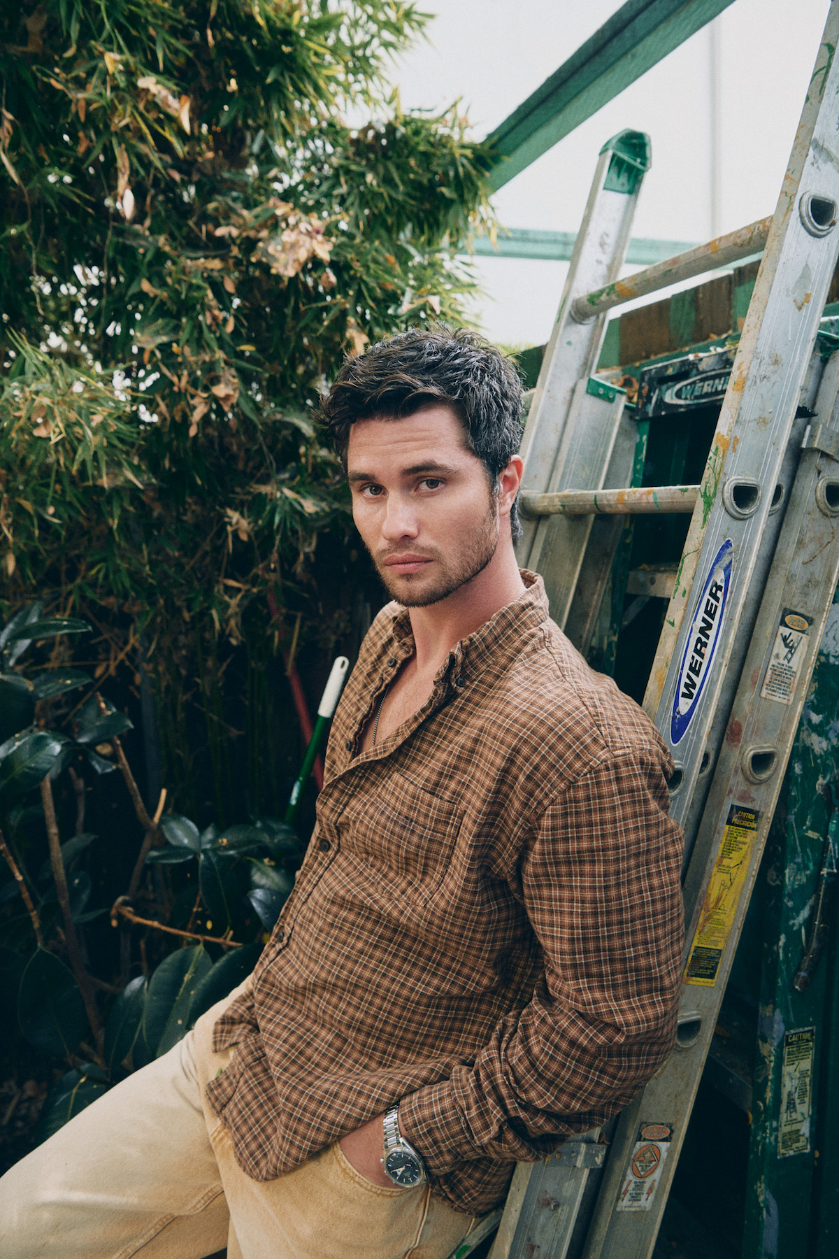 Man in a plaid shirt leaning against a ladder in a garden-like setting with leafy plants and natural light.