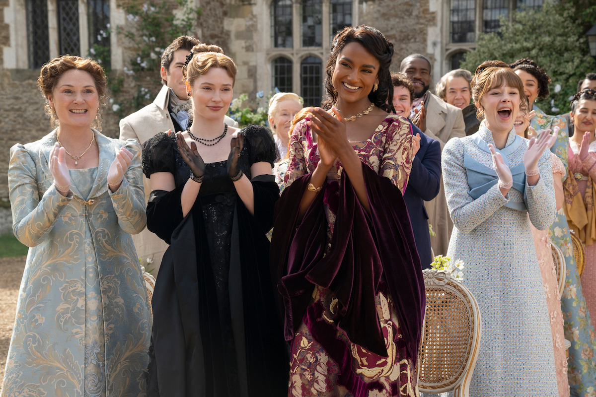 A group of elegantly dressed people in period costumes clap and smile outdoors in front of a historic stone building, suggesting a festive or celebratory event in a garden setting.