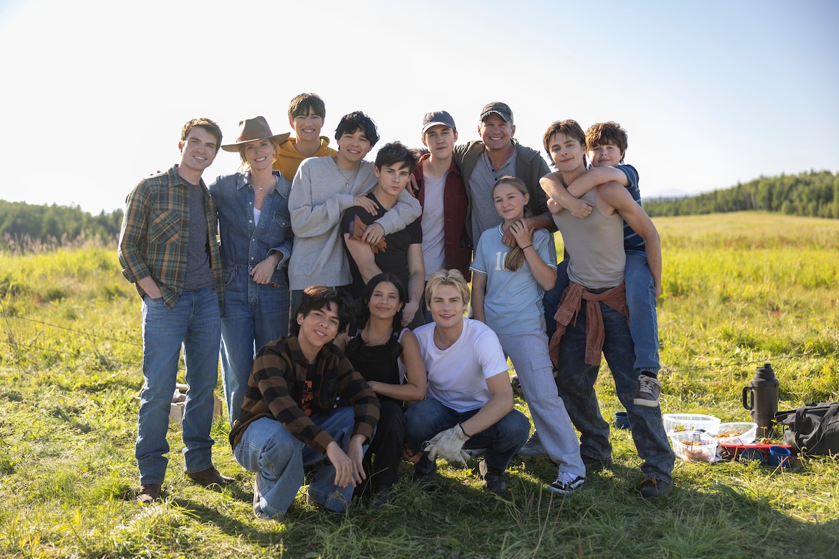 A group of teenagers and adults pose together outdoors on a sunny day in a grassy field, smiling and relaxed, with picnic items on the ground and trees in the background.